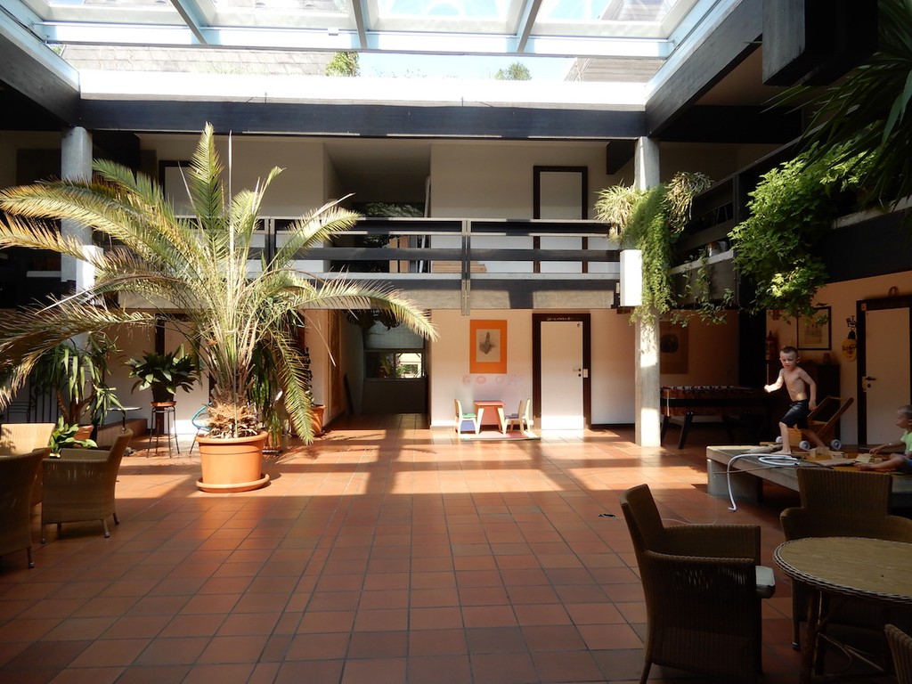 Innenhof mit Sitzgelegenheiten und Palmen. Durch Glasdach strahlt Sonnenlicht.