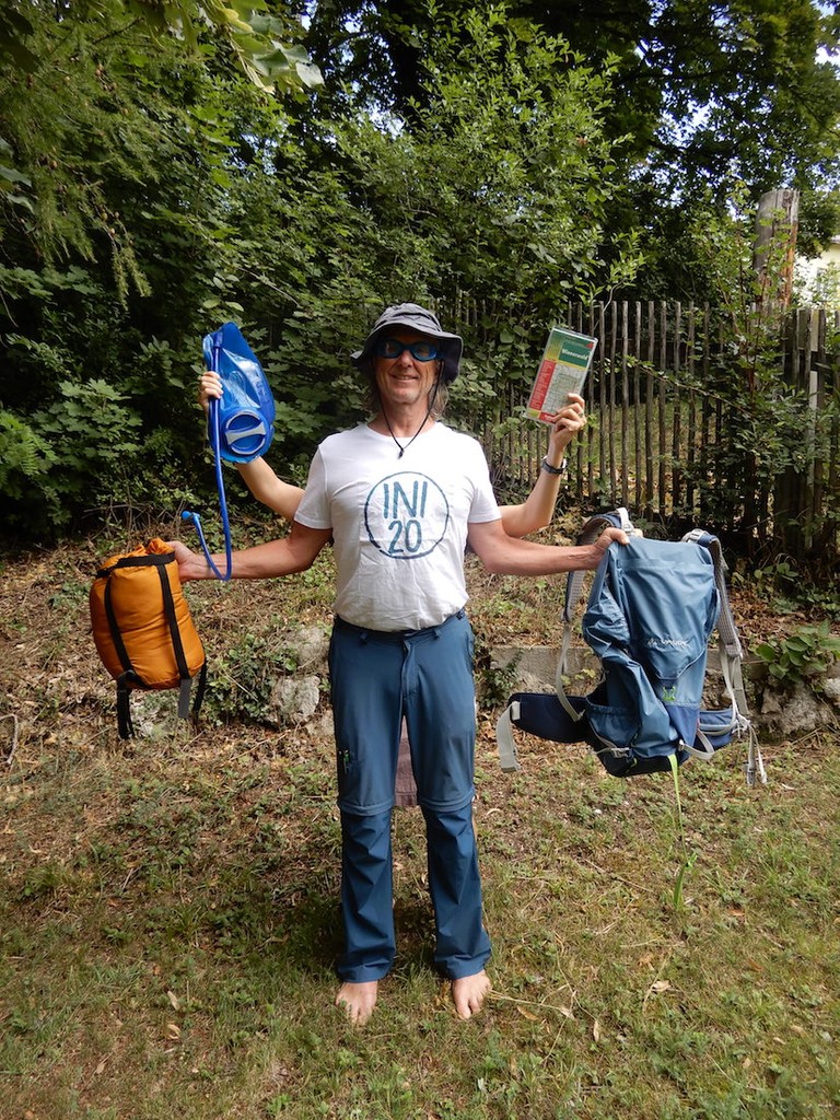 Mann lächelt in die Kamere und hat zwei Rücksäcke in den Händen. Er trägt ein T-Shirt mit der Aufschrift INI20