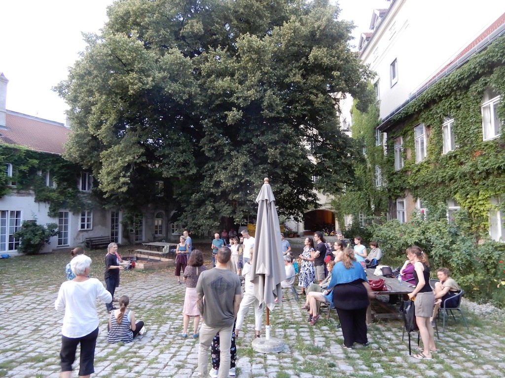 Größere Menschengruppe steht und sitzt in einem Innenhof und hört einer Vortragenden zu